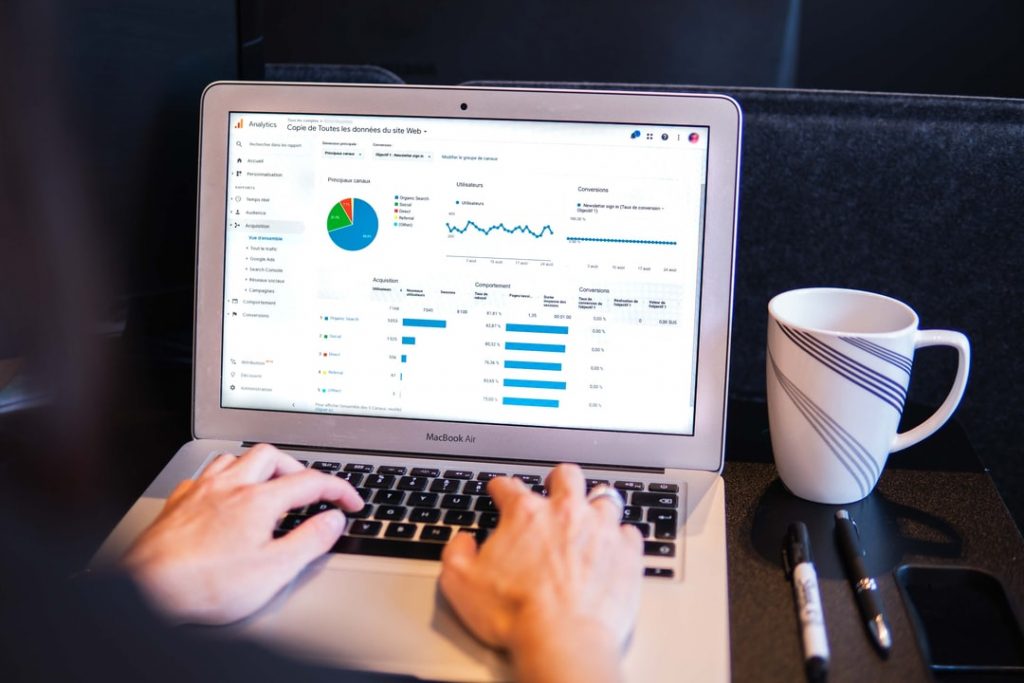 mão masculinas digitando no notebook com uma imagem de dashboard na tela. ao lado seu computador tem uma xícara branca e 2 canetas. O funso da imagem é preto